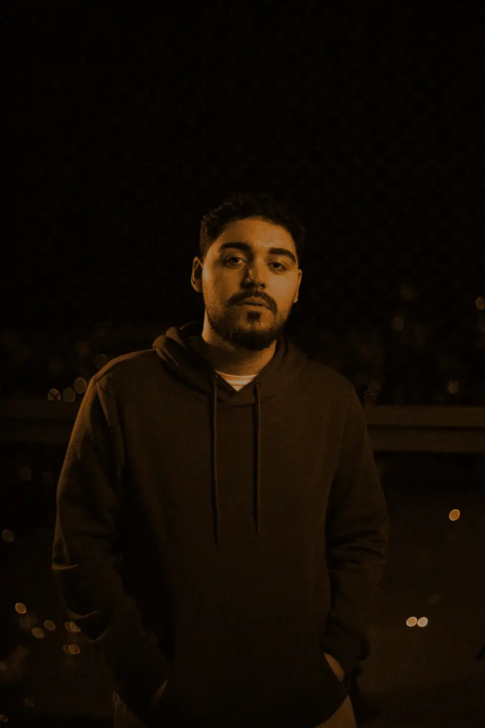 A man in a dark hoodie against a chain-link fence at night